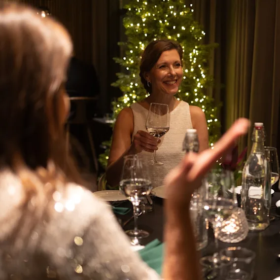 En person smiler og holder et vinglass ved et festlig middagsbord med et pyntet juletre i bakgrunnen.