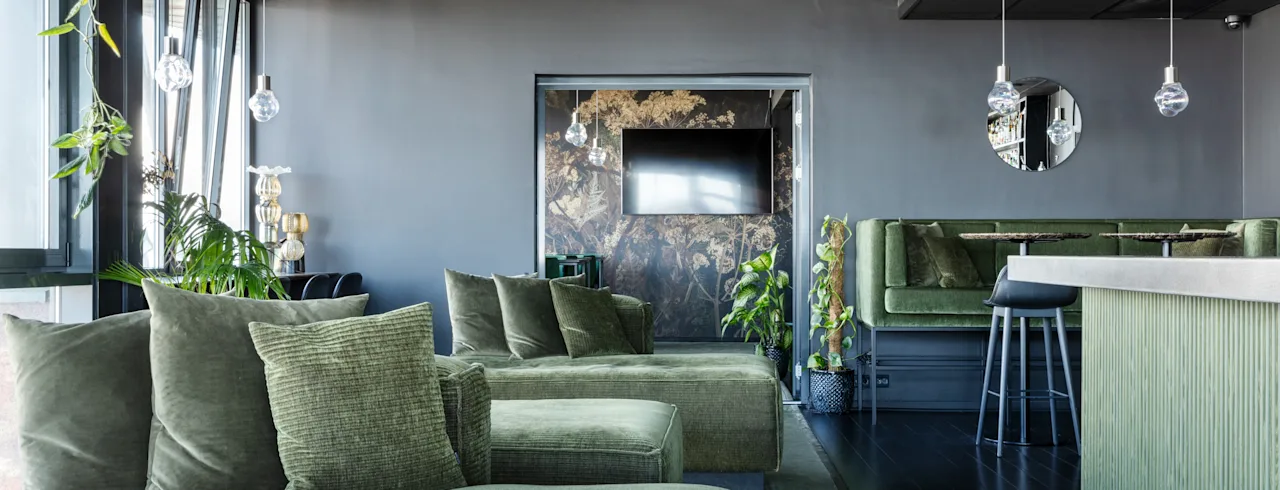 Grønne sofaer står i en elegant, moderne lounge med mørke vegger og gulv. Hengende pendellamper lyser opp rommet, med potteplanter og et speil som skaper en stilfull, avslappet atmosfære.
