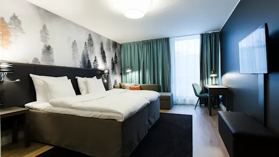 Bed with white linens and pillows, placed in a room with forest-themed wallpaper. Adjacent sofa and desk by a window with teal curtains; wall-mounted TV opposite the bed.