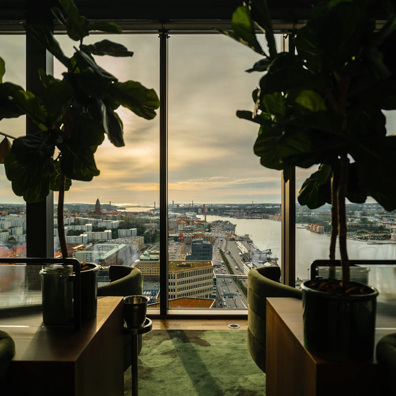 Två krukväxter står på ett bord vid golv-till-tak-fönster med utsikt över en stadsbild med vatten, byggnader och en molnig himmel vid solnedgången.