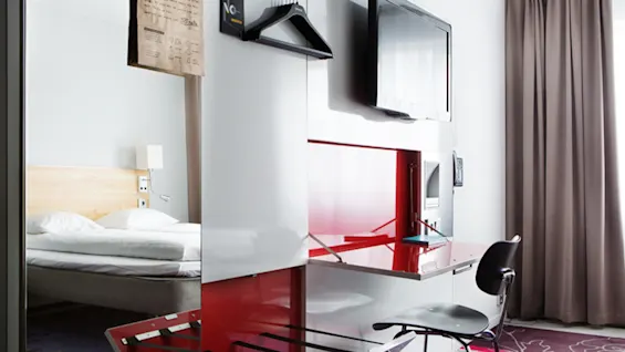 Ett hotellrum har en snyggt bäddad säng, en platt-TV, ett rött skrivbord med en stol och beige gardiner. Klädhängare och bagageställ är synliga i närheten.