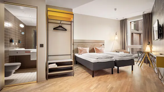 Hotel room with two neatly made beds beside a large window. A modern bathroom with brown tiles is adjacent. The room is furnished with a desk, chair, and hanging rack.