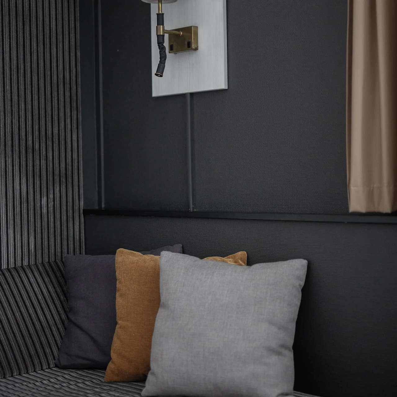 A wall sconce with an angled white shade is mounted above a striped cushioned bench adorned with three colored pillows. Dark walls and a curtain create a cozy interior setting.