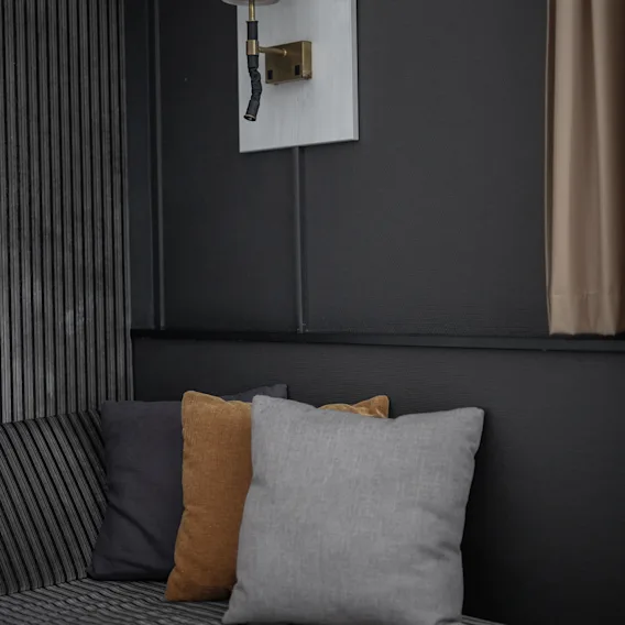 En vägglampa med en vinklad vit skärm är monterad ovanför en randig dyna med tre färgglada kuddar. Mörka väggar och en gardin skapar en mysig interiör.