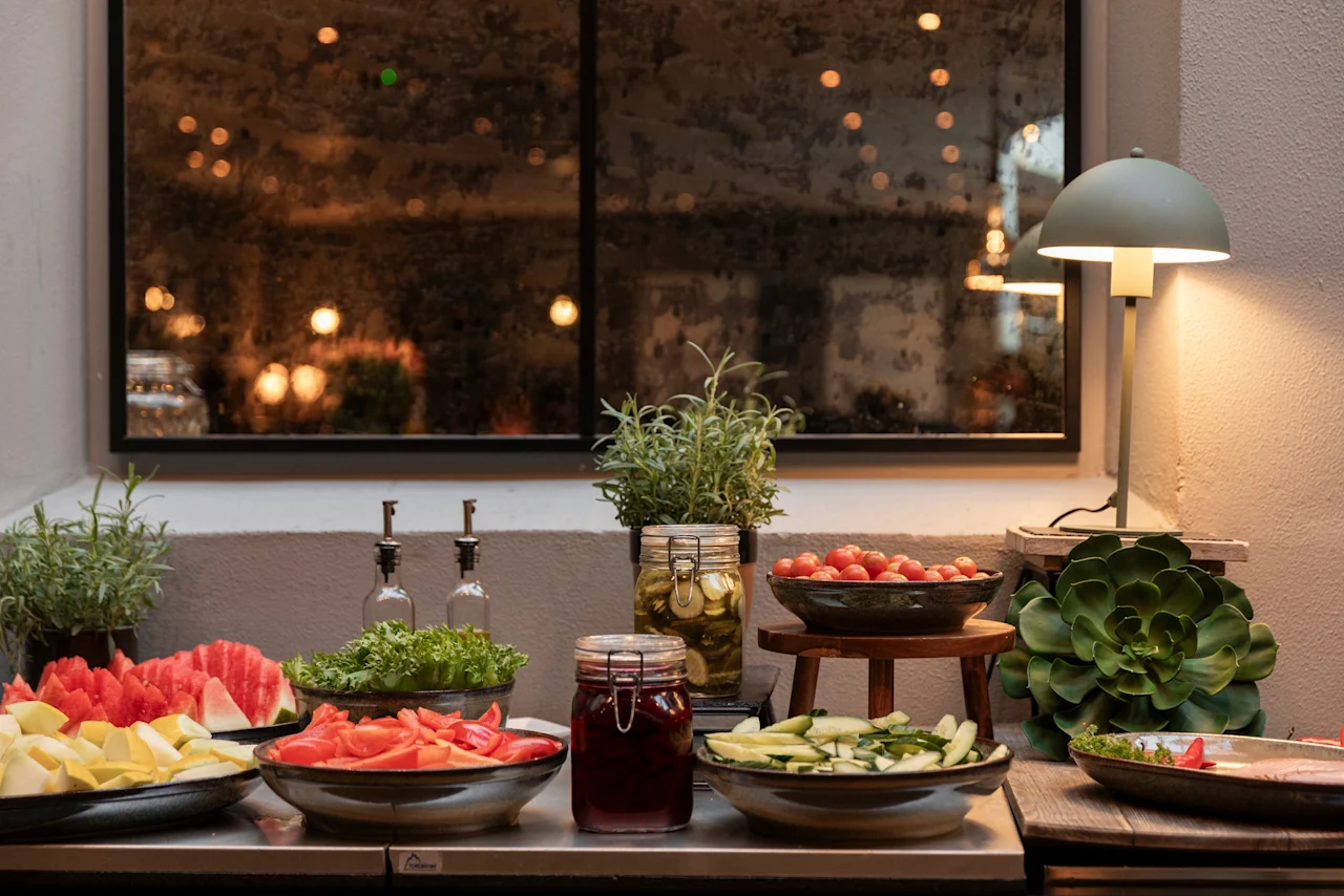A vibrant buffet at Home Hotel Grand Bodø, featuring fresh fruits, vegetables, and pickled items, illuminated by a stylish lamp.
