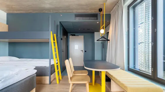 Køjeseng med grå ramme og gul stige i et moderne, minimalistisk værelse. Et bord med træstole og et gult accent står ved et stort vindue med perforeret skærm.