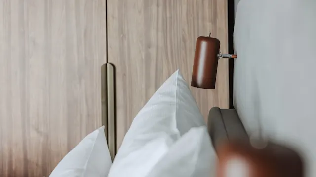 En brun vägglampa är monterad bredvid en säng med vita kuddar, i ett rum med trä garderobsdörrar.
