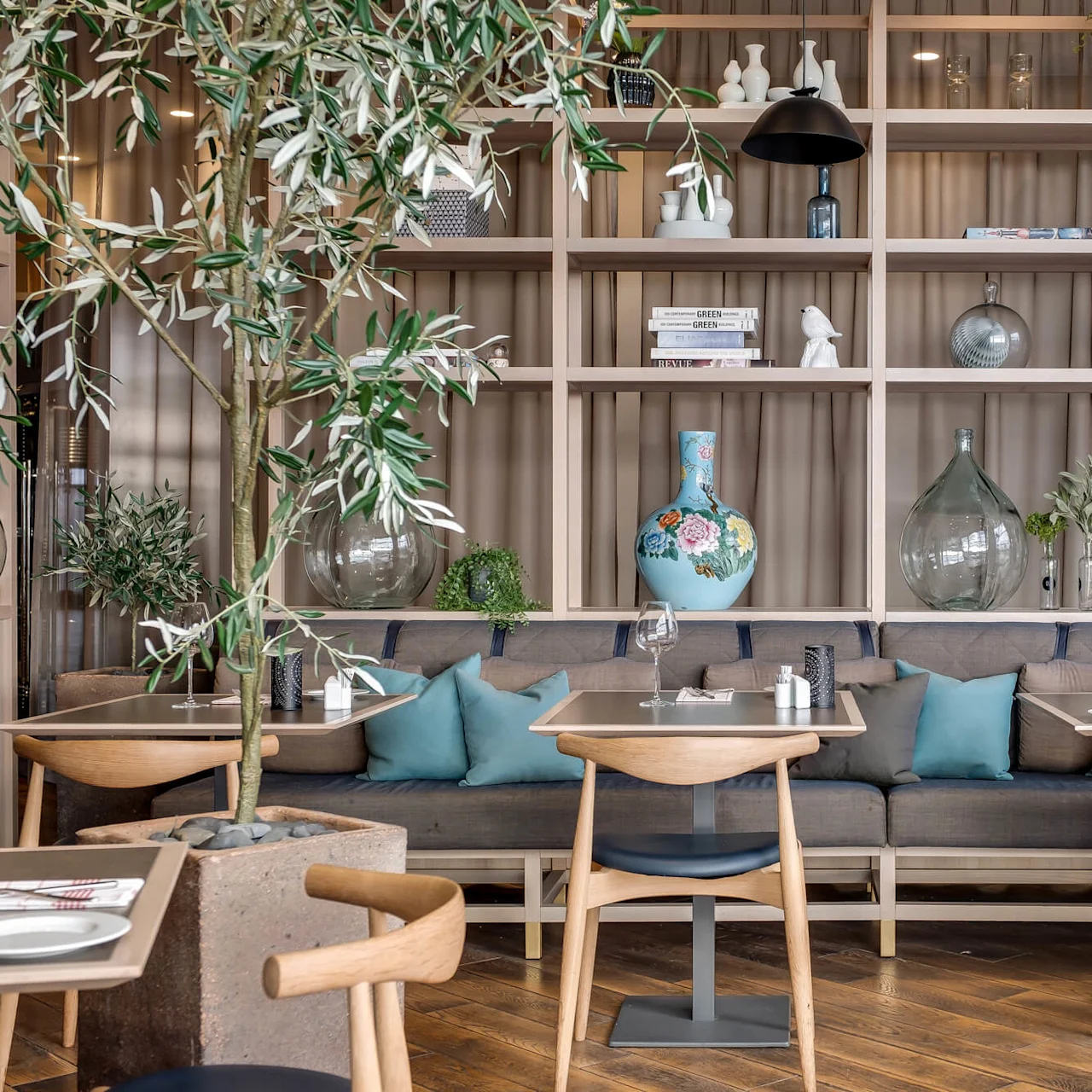 Wooden tables and cushioned benches in a cozy dining area, warmly lit, with decorative shelves and greenery.