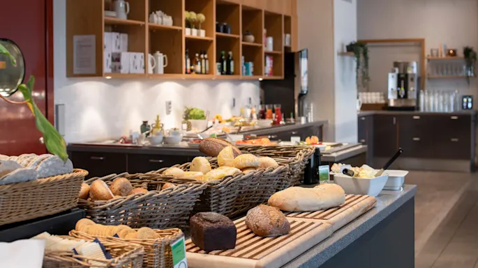 Kurver med brød hviler på en benk, omgitt av ulike frokostvarer i et godt opplyst buffetområde. Hyller med servise og drikke langs veggen.