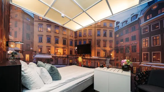 A cozy hotel room features a bed with pillows, a wall mural depicting a picturesque old town scene, and a TV. A small cabinet holds flowers.