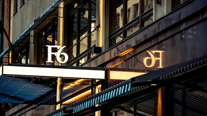 Et skilt med teksten "F6" stikker ut fra en bygningsfasade og kaster en skygge på reflekterende glassvinduer i et bymiljø.