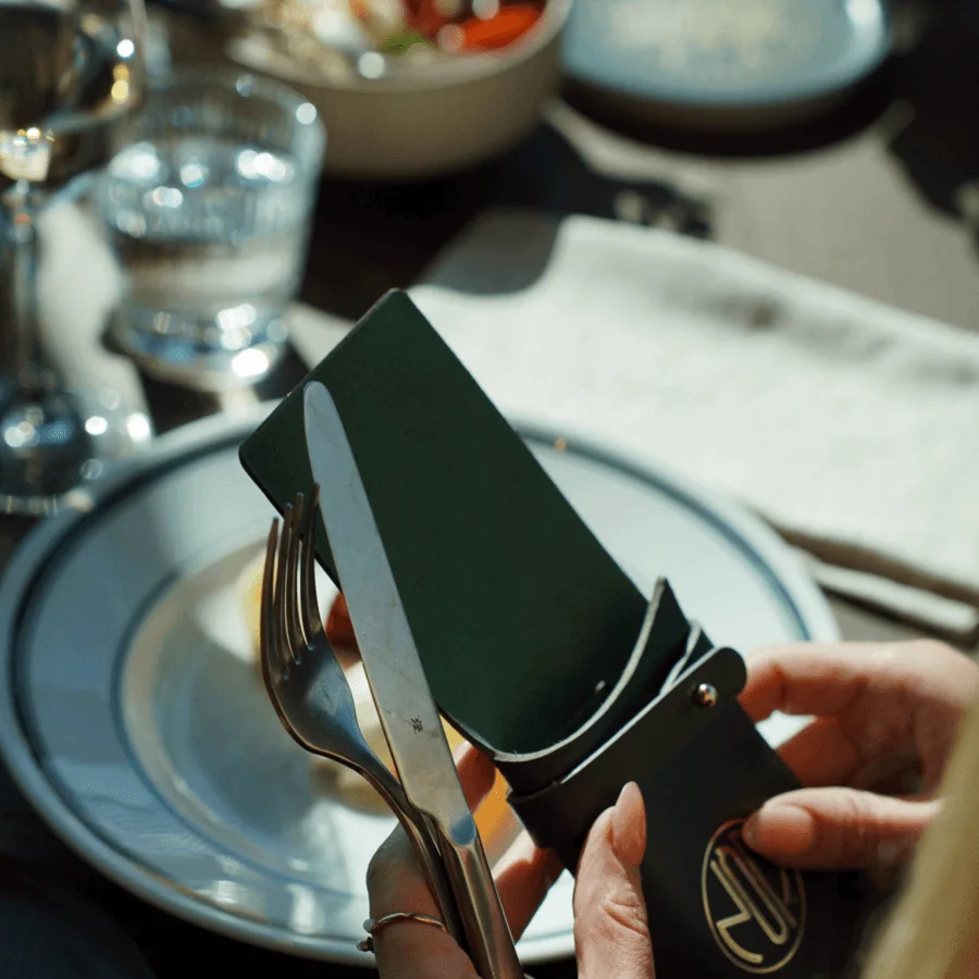 A person holds cutlery, a fork and knife, in a leather case near a dining table with plates and glassware, creating an elegant dining setting.