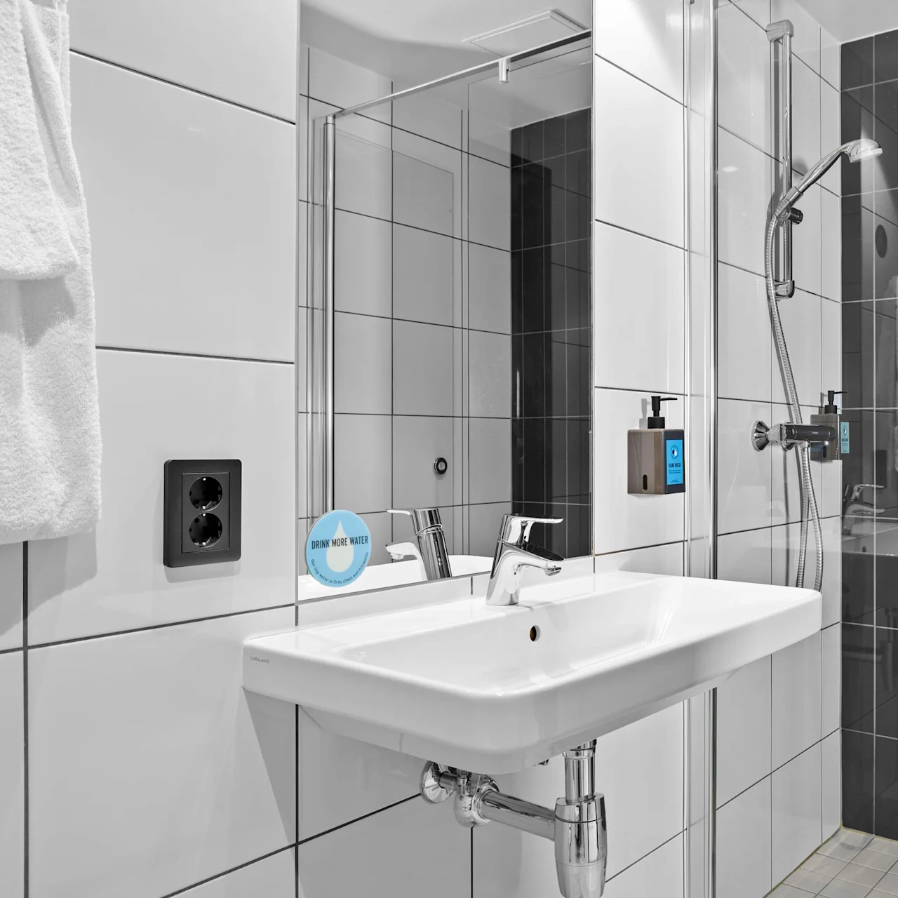 A white sink stands with a chrome faucet in a modern bathroom, surrounded by white and black tiled walls. A sticker on the mirror reads, "Drink More Water."
