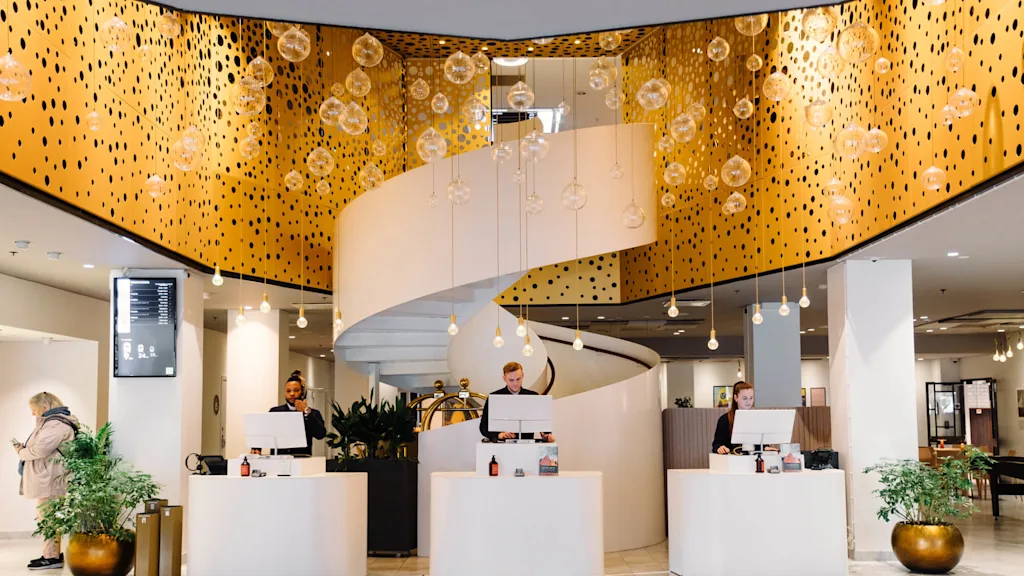 Reception med tre personer bakom moderna, vita skrivbord under ett dekorativt, gyllene tak med hängande glasglober. En spiraltrappa stiger i bakgrunden. Växter pryder rummet.