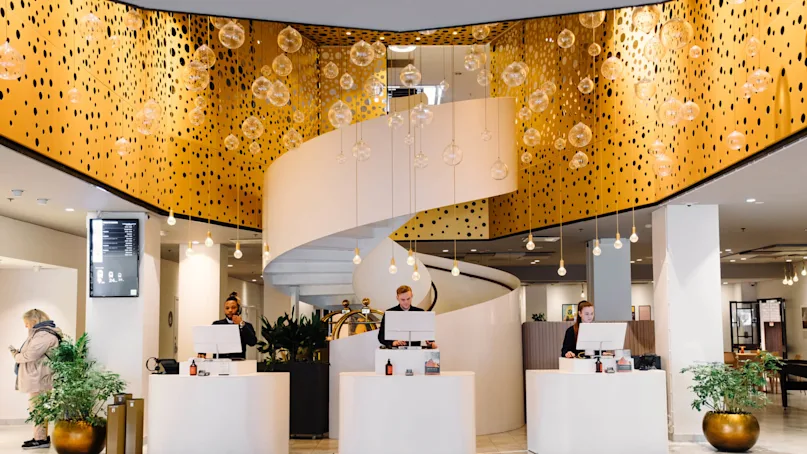 Reception area with three people behind modern, white desks under a decorative, golden ceiling with hanging glass orbs. A spiral staircase ascends in the background. Plants adorn the space.