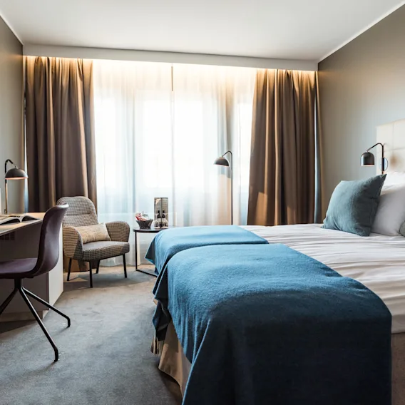 Ett prydligt hotellrum har en stor säng med blå täcken och kuddar, en stol och ett skrivbord med lampa, mot ett fönster med gardiner.