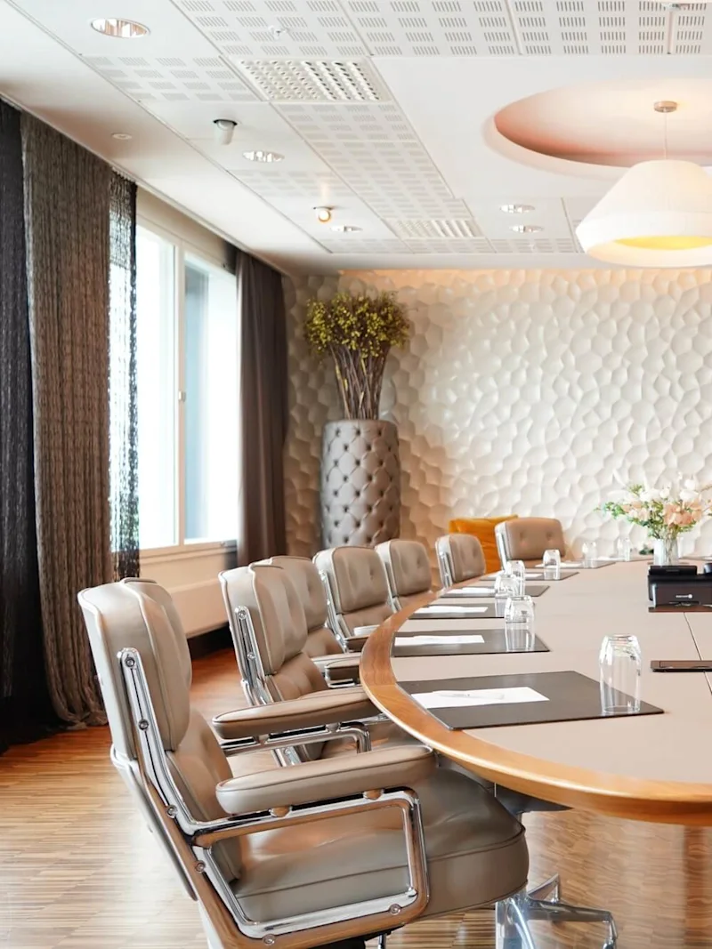 A large oval conference table surrounded by leather chairs, set in a modern room with textured walls, large planters, and windows with curtains. Papers and glasses are on the table.
