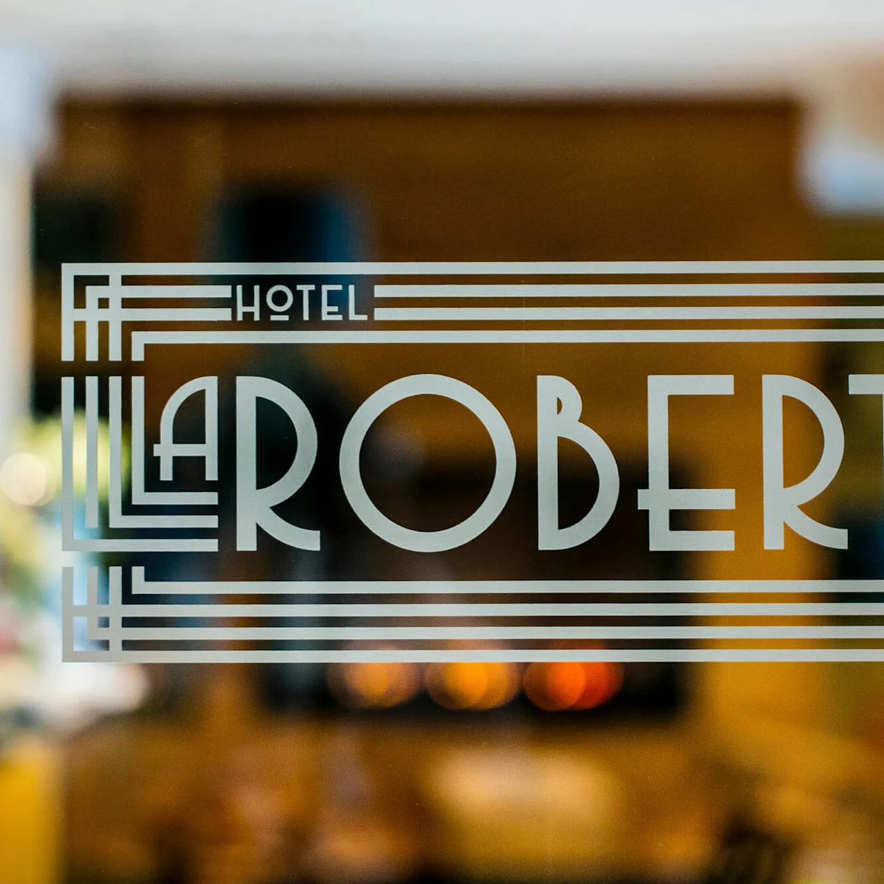 Glassrute viser hvit, art deco-stil tekst "HOTEL ROBERTS" med geometriske mønstre; dempet opplyst interiør med uklar, varm atmosfære synlig bak glasset.
