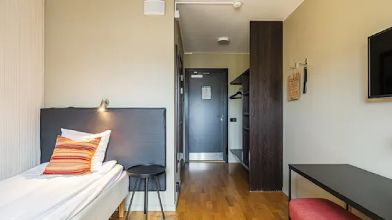 Et koselig hotellrom med enkel innredning: en seng med en fargerik pute, et lite rundt bord, åpent garderobeskap og en TV montert på veggen. Atmosfæren er varm og innbydende, perfekt for avslapning.