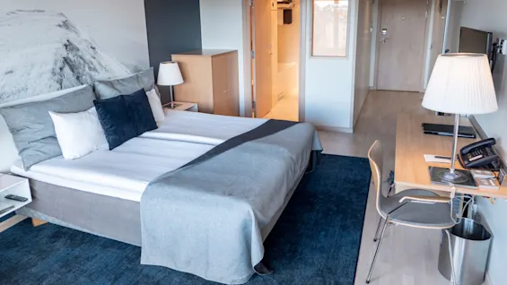 Ett prydligt hotellrum innehåller en säng med kuddar och en grå filt. Bredvid sängen står ett skrivbord med en lampa och en stol mot väggen. En dörr leder till ett ljust badrum.