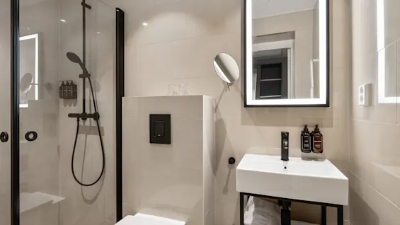 Et moderne og rent hotelbadeværelse med bruser, toilet og en vask med et oplyst spejl.