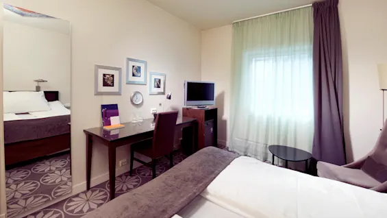 A neatly arranged hotel room with a bed, desk, TV, and sitting area, bathed in natural light from a curtained window.