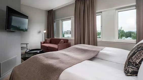 Seng med beige teppe, ved siden av en maroon sofa og veggmontert TV, i et lyst rom med store vinduer og gardiner; en gulvlampe står i nærheten.