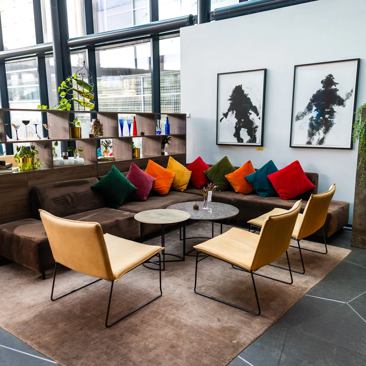 Et siddeområde har en brun sofa med farverige puder og fire beige stole omkring runde borde. Hylder med planter og dekorationer skaber en hyggelig atmosfære, sammen med abstrakt kunst på hvide vægge.