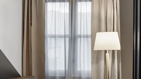 Et hyggeligt hotelværelseshjørne med en lænestol, et lille bord, en gulvlampe, et vindue med gardiner og en glitrende lysekrone.