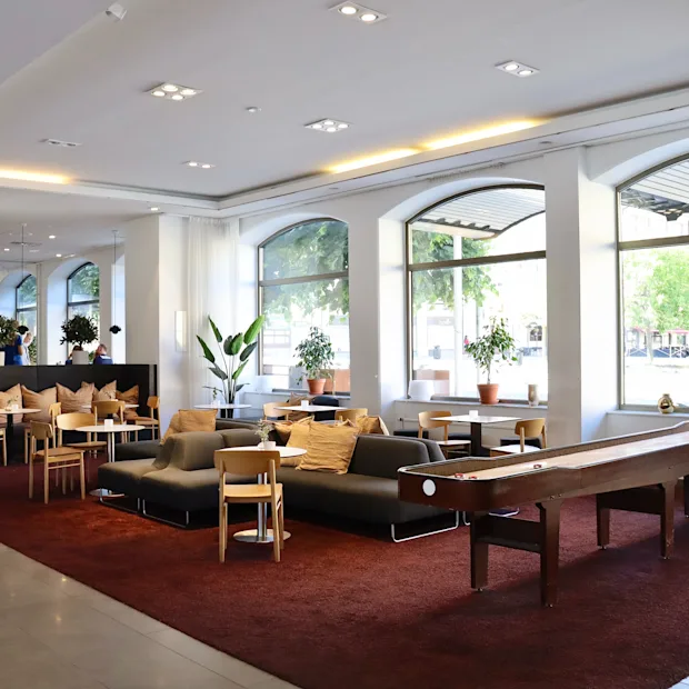 Norsk bokmål: Loungeområde med sofaer og stoler på et rødt teppe, med planter og store vinduer mot gaten. Et shuffleboardbord kompletterer det moderne, koselige interiøret.