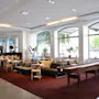 Lounge area featuring sofas and chairs on a red carpet, with plants and large windows overlooking the street. A shuffleboard table completes the modern, cozy interior.