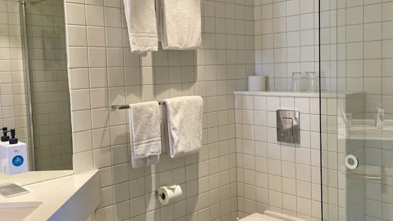 Et moderne bad har en vegghengt toalett, flislagte vegger og en dusj med glassvegg. Håndklær henger på stenger nær en toalettrull. En servantskap med såpe er synlig.