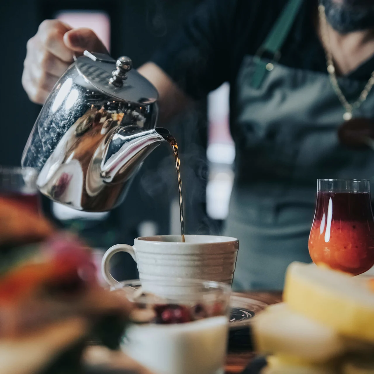 En person häller kaffe från en silverkanna i en vit mugg på ett träbord, omgivet av smörgåsar, ett glas juice och en leende man i förkläde.