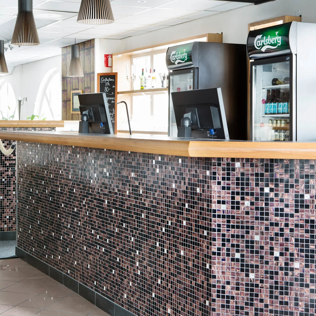 A mosaic-tiled bar counter stands empty in a clean, well-lit restaurant with tables set for dining in the background.
