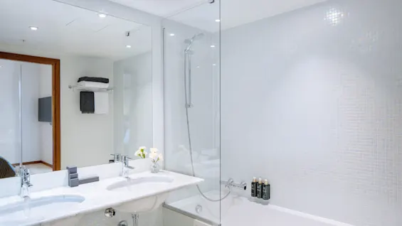 Lyst, moderne badeværelse med en hvid håndvask og dobbelt vaske. En glasskærm adskiller badekarret, fyldt med toiletartikler. Hvide fliser og et håndklædestativ forstærker det rene, minimalistiske design.