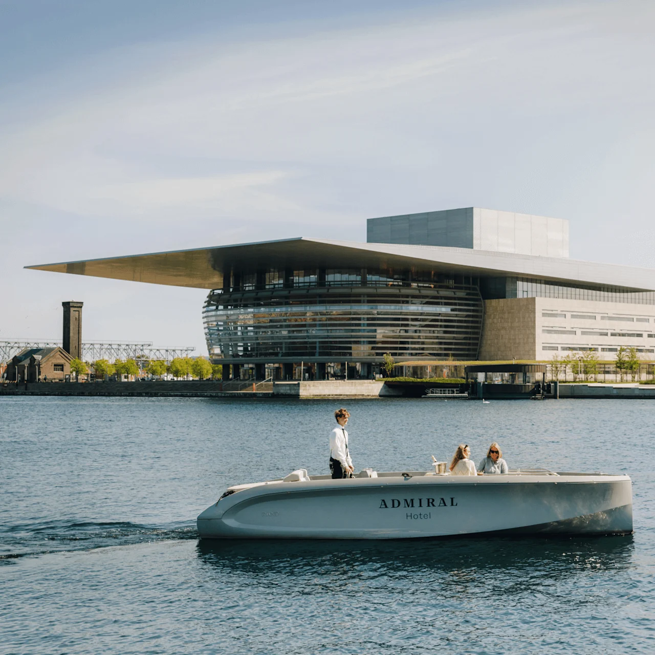 Boat marked "Admiral Hotel" cruises with three people; one steering, two seated. Surrounding is a harbor featuring a large modern building with tiered design and expansive roof.