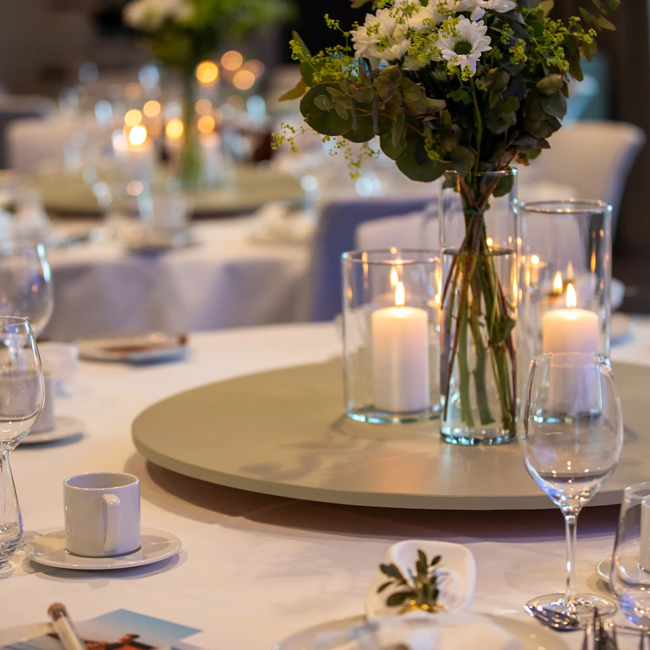 Et rundt spisebord er elegant dekket med hvit duk, med vaser med blomster og lys på et stort rundt fat. Glass og tallerkener er pent ordnet i en mykt opplyst bankettsal.