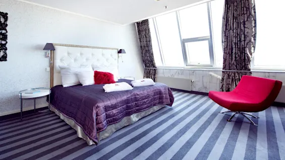 A well-lit bedroom with a large bed and a striking red chair, against a backdrop of windows draped in patterned curtains, over a striped carpet.