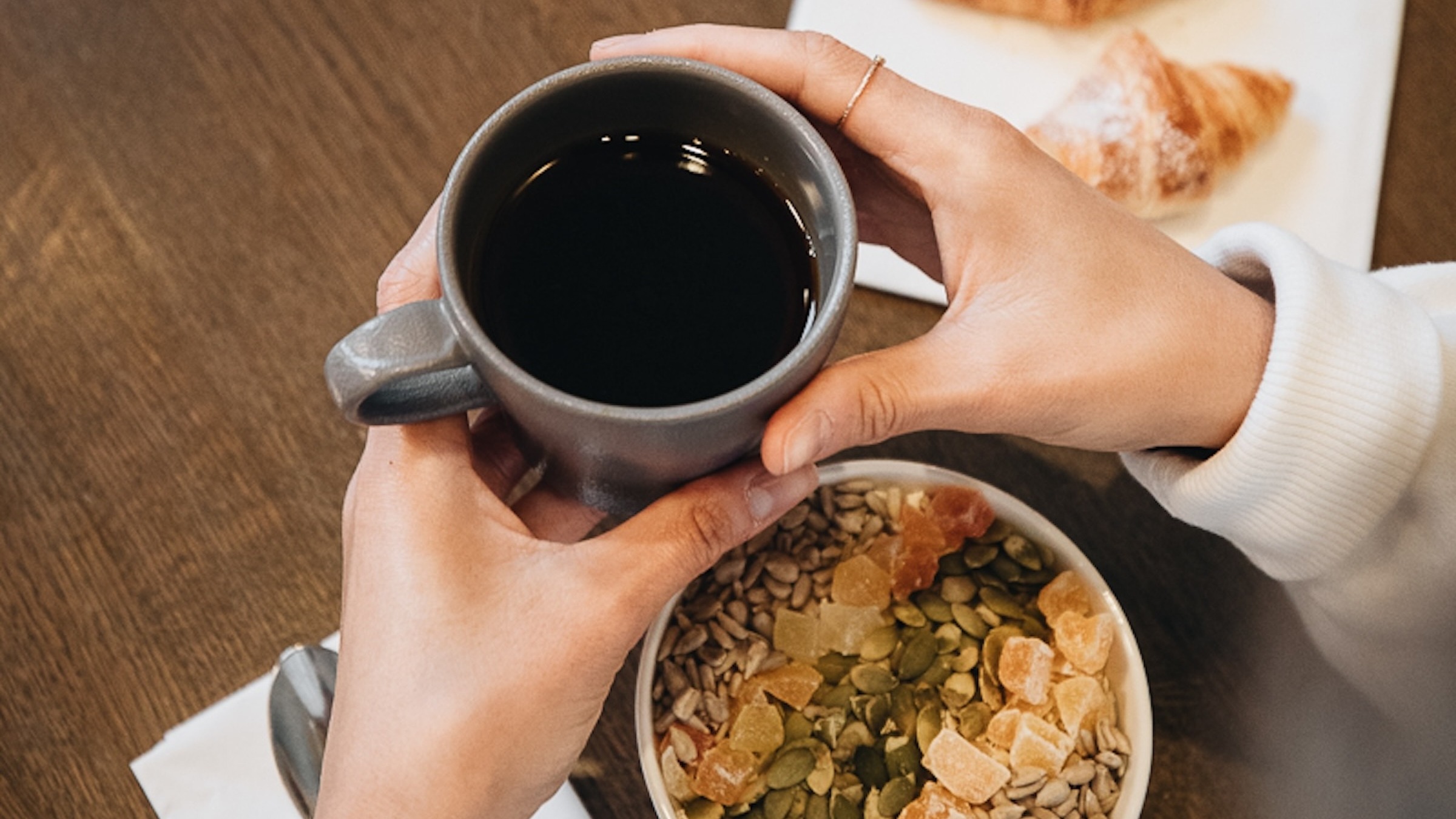 A person holding a coffee cup, with a healthy breakfast bowl and croissants on a wooden table.