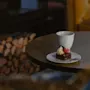 En liten kaka med grädde och bär ligger på en tallrik bredvid en kopp på ett runt bord; trästockar är staplade i bakgrunden nära en mjuk stol.