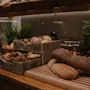 Brödlimpor och bullar på en träbänk med skärbrädor; krukväxter och en brödrost i bakgrunden skapar en varm, rustik köksmiljö.
