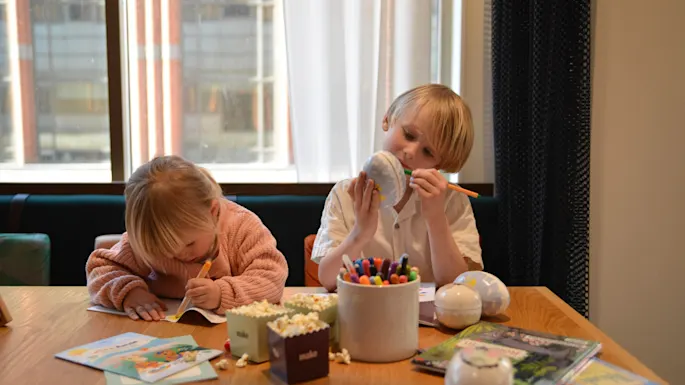 Two children engaged in drawing and coloring, seated at a wooden table cluttered with markers, books, and popcorn under a large window.
