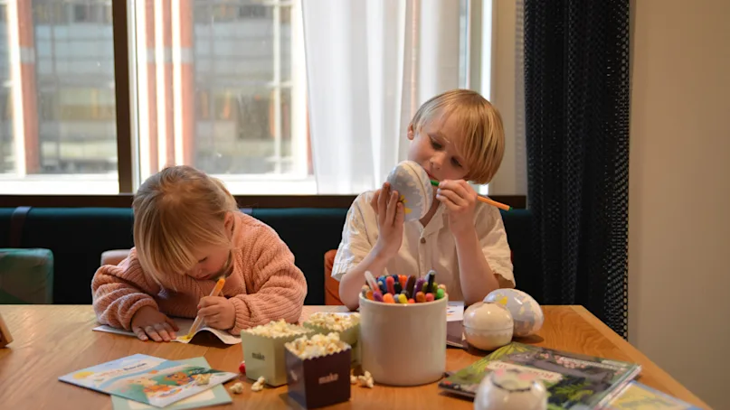 Two children engaged in drawing and coloring, seated at a wooden table cluttered with markers, books, and popcorn under a large window.