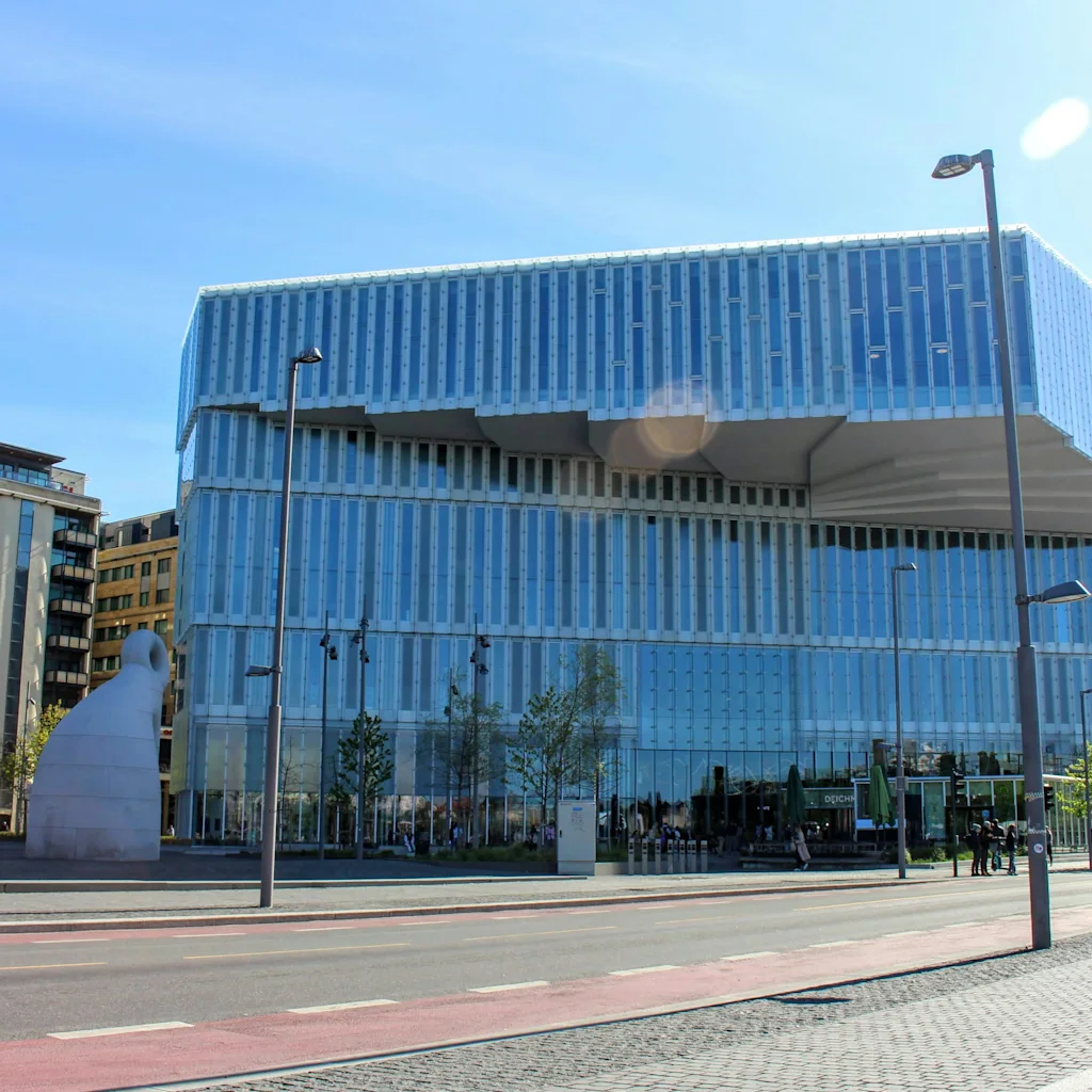 En moderne glassbygning med unik design en solrik dag, med folk som går langs gaten i forgrunnen.