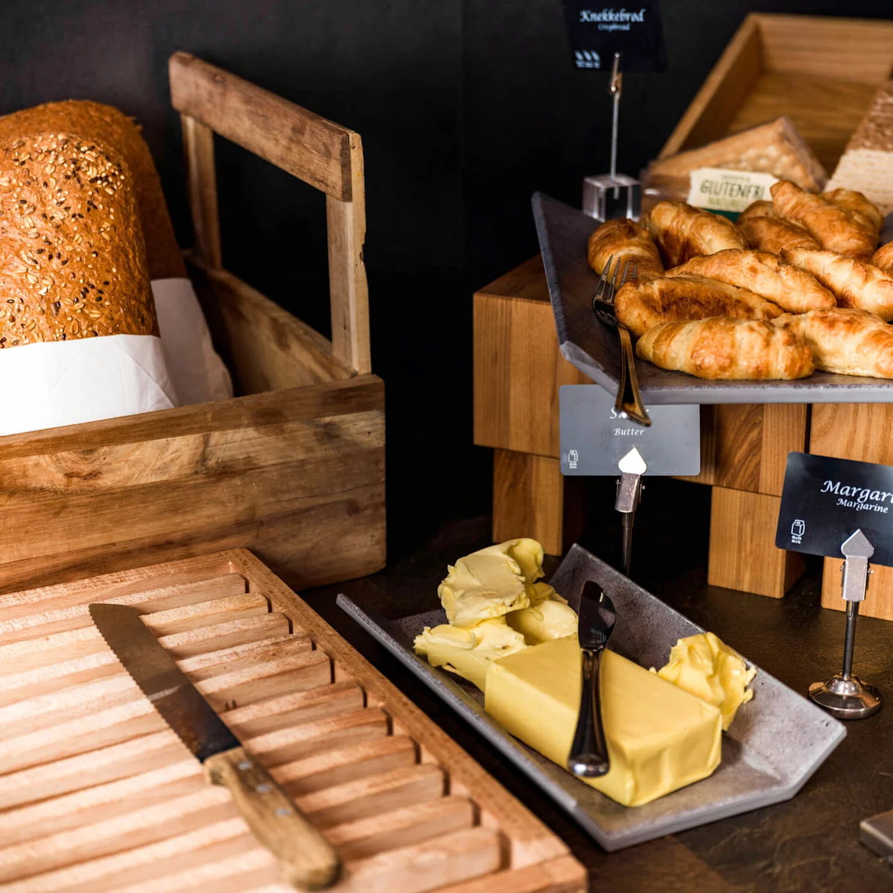 Bread slices sit in a wooden box next to a tray of croissants. Butter and margarine are labeled on a stone dish beside a cutting board with a knife.