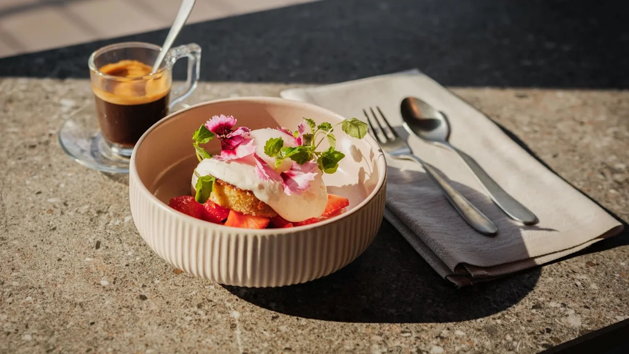 En skål med efterrätt med vispad grädde, jordgubbar och ätbara blommor står på ett stenbord bredvid en espressokopp och bestick på en servett.