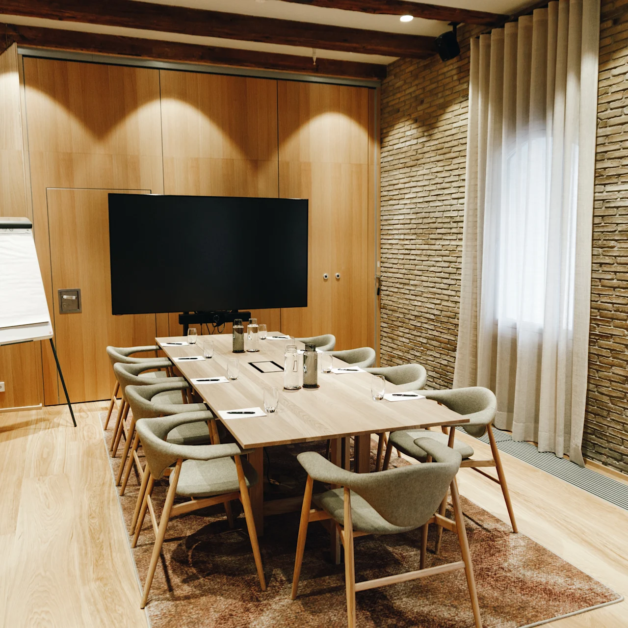 A conference table equipped with chairs, paper, and pens is set in a modern meeting room. A large screen is mounted on the wall; a flip chart stands nearby.