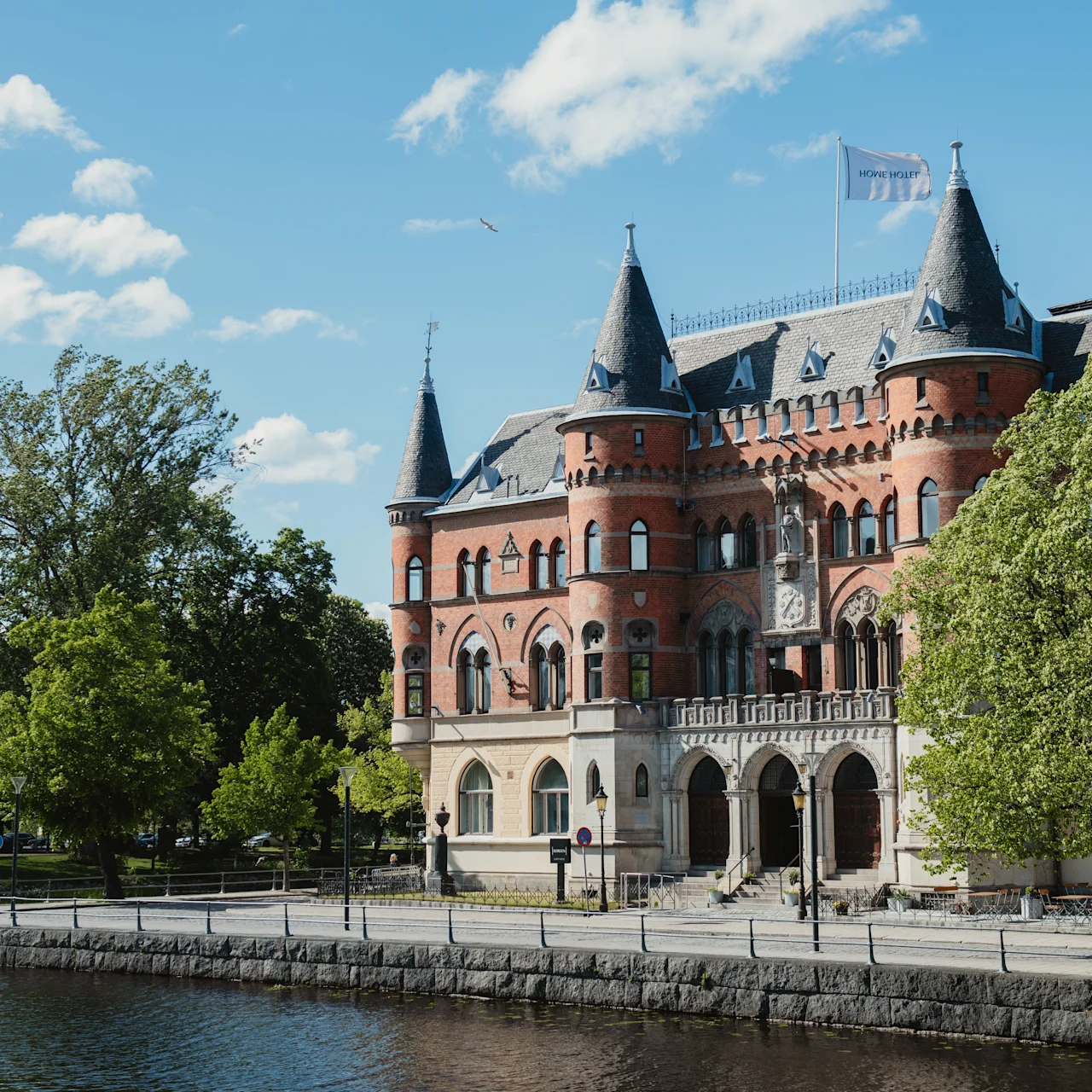 En stor röd tegelstensbyggnad liknande ett slott med spetsiga torn står fridfullt vid en flodpromenad, omgivet av frodiga gröna träd under en klarblå himmel.