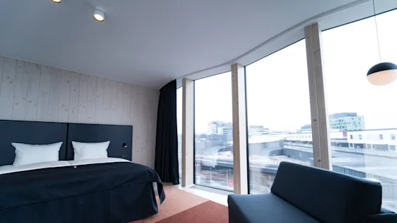 A bed with white pillows and a dark duvet sits in a modern room, adjacent to a large window showing a cityscape. A sofa and lamp are nearby.
