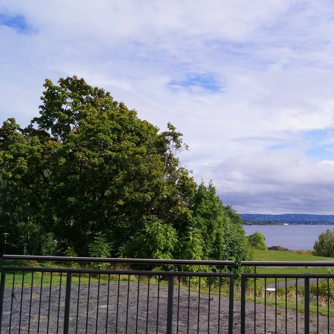 Railings frame a scenic view with lush trees and a grassy hill leading down to a calm lake. The sky is partly cloudy with patches of blue.